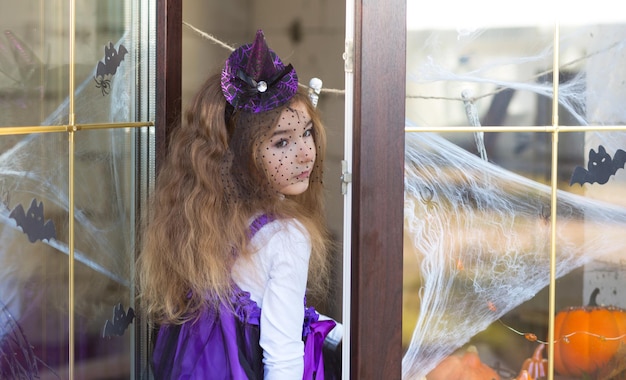 A girl in a witch costume on the windowsill by the window is playing with a spider web and a spider decorating the house for the Halloween holiday Terrible scenery fear and horror pumpkins