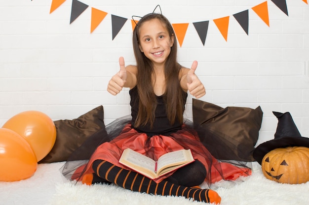 girl in a witch costume with a witchcraft book celebrating halloween at home shows her fingers cool