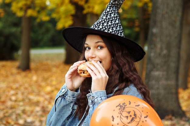 a girl in a witch hat next to an orange balloon for Halloween eats a burger in an autumn park