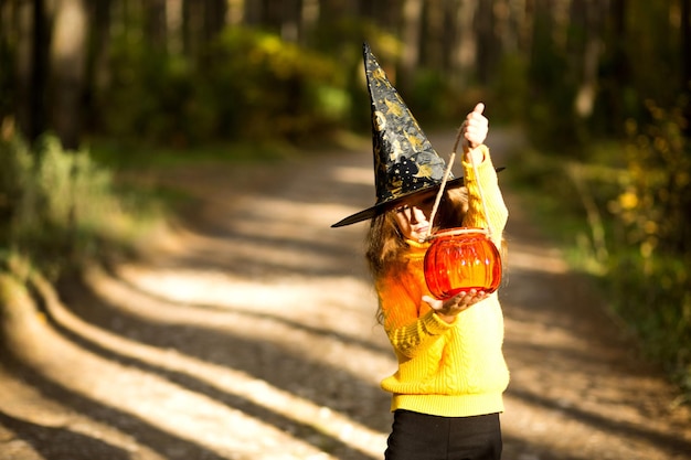 A girl in a witch's hat in a yellow sweater and with a pumpkin a lantern and a bucket for sweets in autumn forest Halloween Holiday Portrait closeup