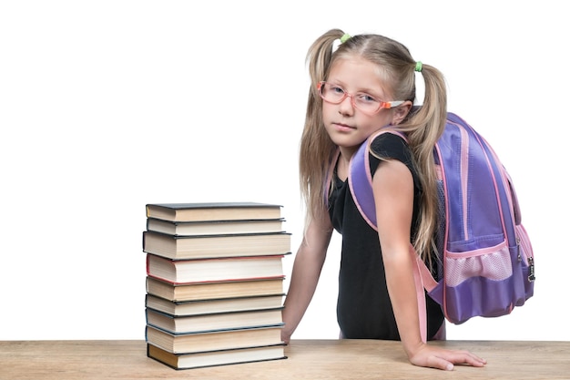 Girl with a backpack in glasses leaned against a wooden table with a stack of books isolated on a white background