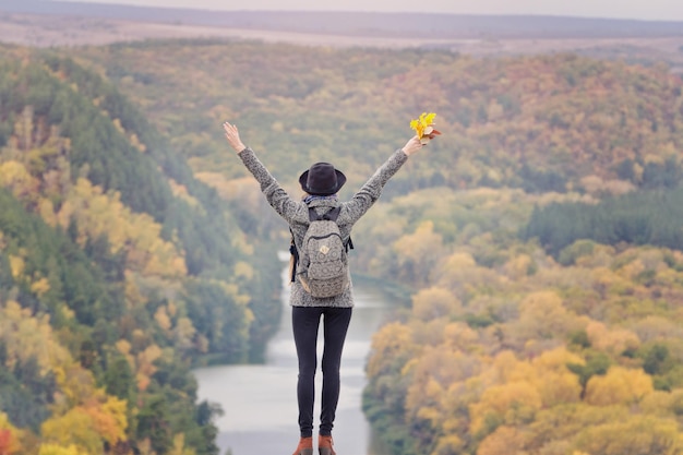 Girl with a backpack and a hat standing on a hill Hands raised up River and mountains below Back view