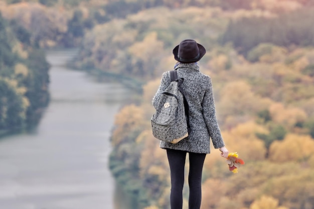 Girl with a backpack and a hat standing on a hill River and forest below Back view