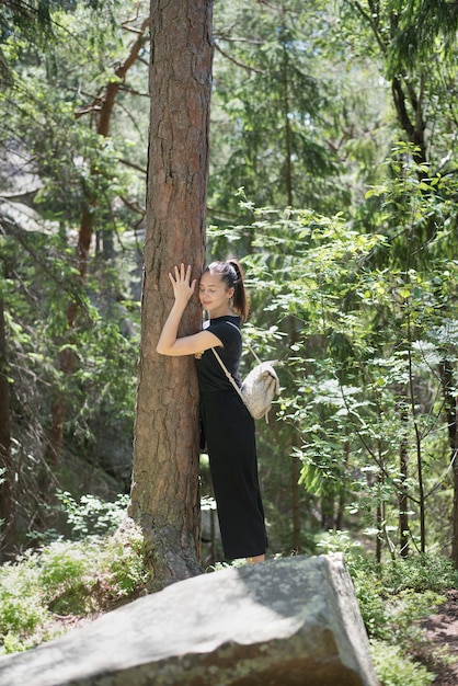 Girl with a backpack is hugging a tree with her eyes closed in the forest. Summer sunny day.