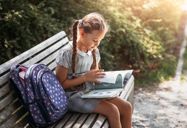 Girl with a backpack sitting on a bench and reading a book near the school Back to school lesson schedule a diary with grades Education primary school classes Back to school