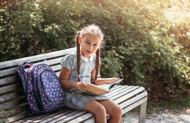 Girl with a backpack sitting on a bench and reading a book near the school Back to school lesson schedule a diary with grades Education primary school classes Back to school