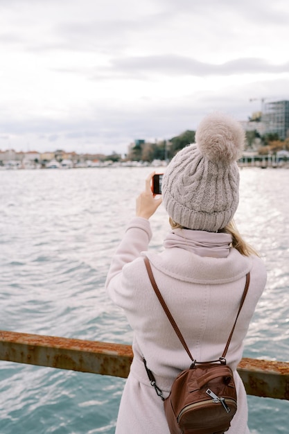 Girl with a backpack stands on the pier and takes pictures of the sea coast back view