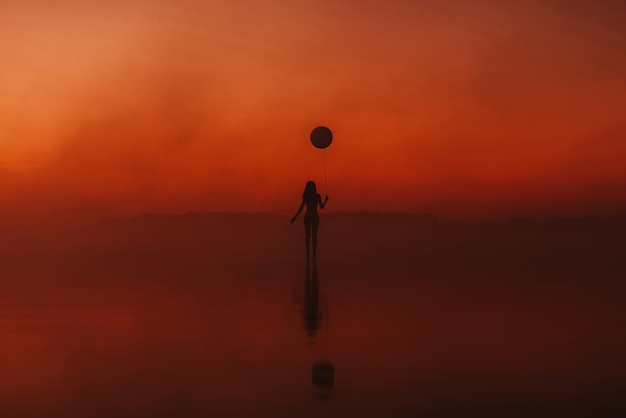 Girl with a balloon in her hand on the water at sunrise in the fog in summer