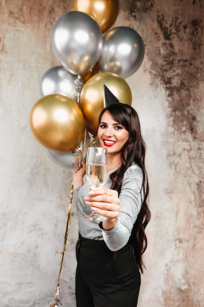 A girl with balloons is laughing against the background of a decorative wall A beautiful happy woman on the birthday of a New Year's holiday party having fun holding a glass of champagne