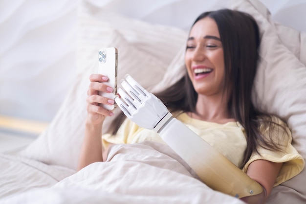 Girl with bionic arm using phone reading social media and chatting in bed woman with disability learning or working online remotely from home