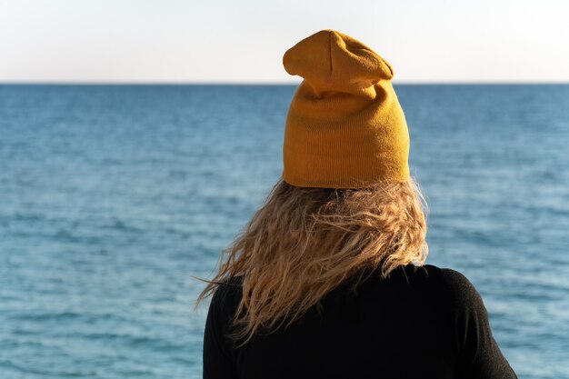 A girl with blond curly hair in black sport jacket and yellow cap is looking at the blue sea