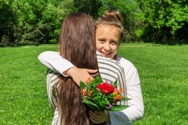 A girl with blond hair combed in a bun gives her mother a bouquet of red flowers