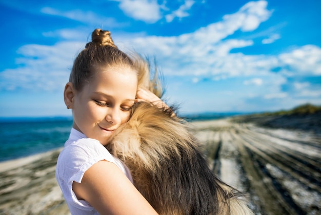 Girl with blond hair with smile hugs pomeranian dog with golden hair on seashore near Black Sea