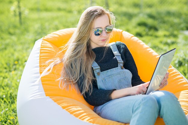 Girl with blond long hair with a tablet sitting on an inflatable chair on a green lawn