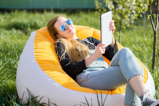 Girl with blond long hair with a tablet sitting on an inflatable chair on a green lawn