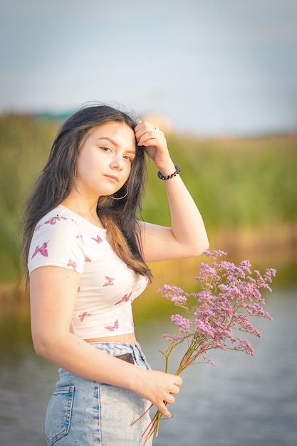 A girl, with a bouquet of flowers in her hand, fixes her long dark hair, at sunset near a lake.