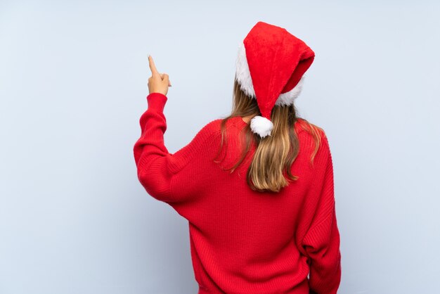 Girl with christmas hat over isolated blue background pointing back with the index finger