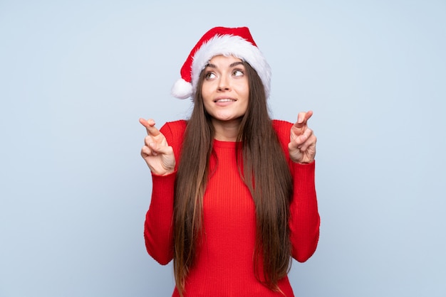 Girl with christmas hat over isolated blue with fingers crossing