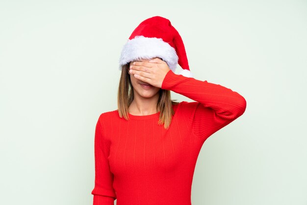 Girl with christmas hat over isolated green covering eyes by hands