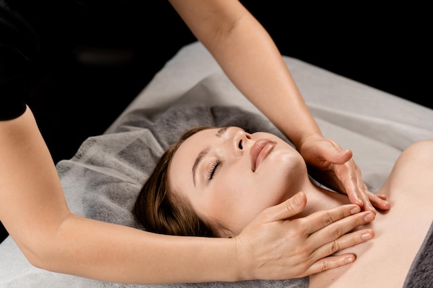 Girl with closed eyes is relaxing on face and neck massage in spa Masseur is making facial massage for young woman in spa Facebuilding and relaxing procedures