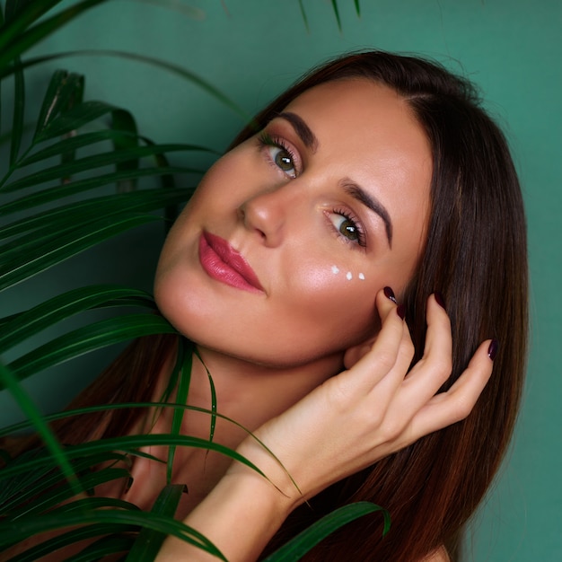 Girl with cream dots on her face between palm leaves on green background