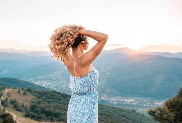 A girl with curly hair in summer dress on a background of mountains at sunset