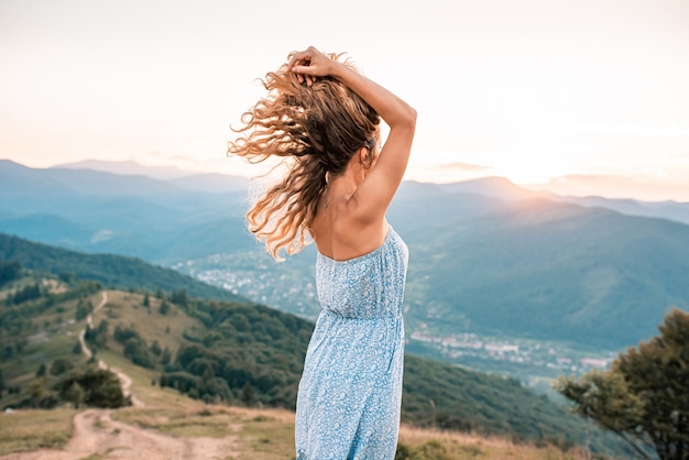 A girl with curly hair in summer dress on a background of mountains at sunset