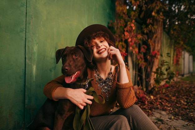 girl with curly hair and vintage clothes sits on the ground near a country house hugs the dog