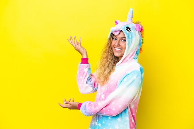 Girl with curly hair wearing a unicorn pajama isolated on yellow background extending hands to the side for inviting to come