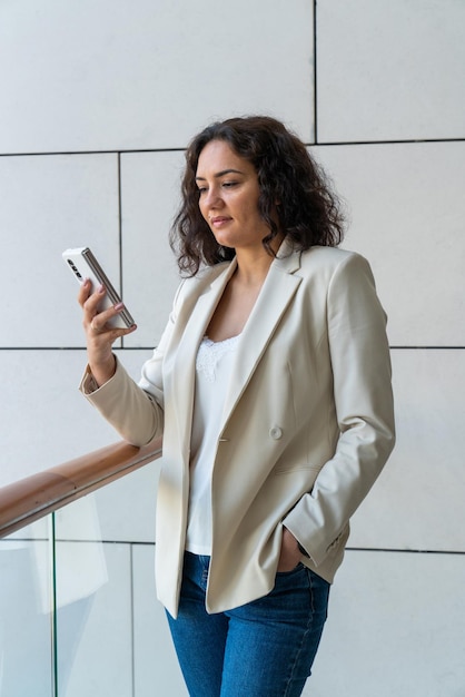 A girl with dark hair stands near a large window against the wall and looks into a smartphone