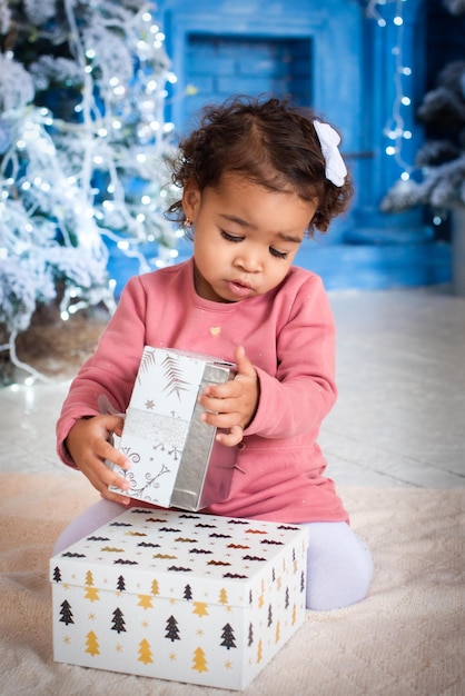 Girl with dark skin color lies under the Christmas tree. cute mulatto is celebrating the new year. African girl unwraps gifts. garland, lights, bokeh.