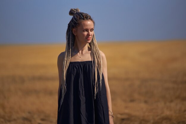 A girl with dreadlocks in a long black dress stands in the middle of a field with wheat and looks aw...