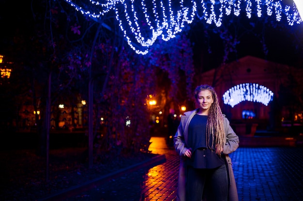 Girl with dreadlocks walking at night street of city against garland lights.