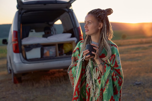 A girl with dreadlocks wrapped in a bright blanket holding a cup in her hand standing in a field at ...