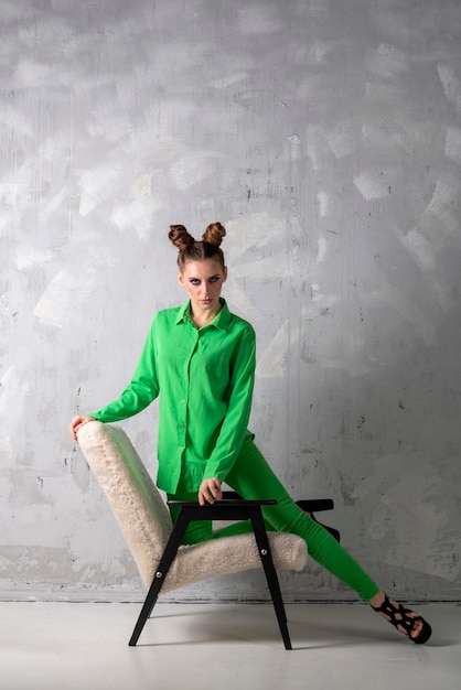 Girl with evening makeup and bun hairstyle poses leaning on chair in modern studioYoung woman in fashionable green suit