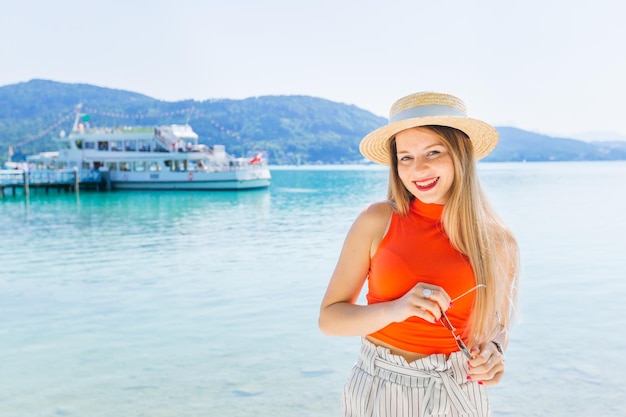 A girl with freckles in a boater and holds sunglasses in her hand a lake a pier