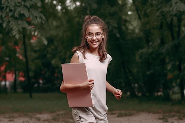 A girl with glasses is running in a park and holding a pink notebook.