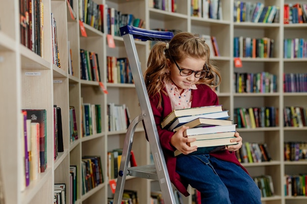 Girl with glasses reads a book sitting on a stepladder in the library