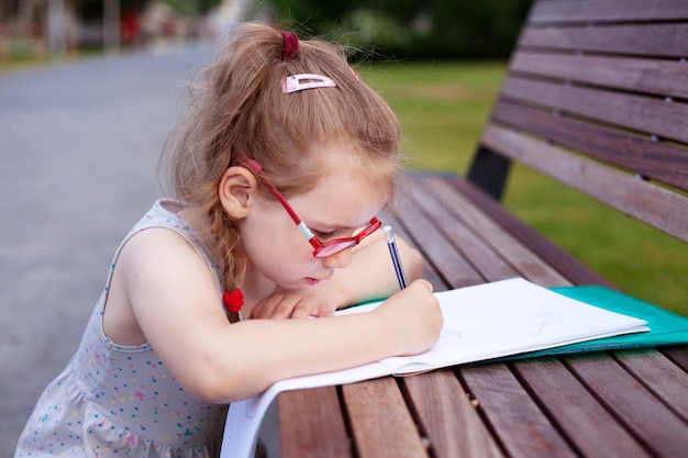 Girl with glasses writes and draws in a notebook outdoors. homework in nature.