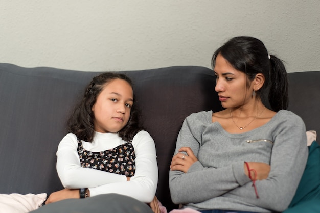 Girl with her arms crossed looking at the camera while her mother looks at her angrily. Mother and daughter sitting on the sofa annoyed.