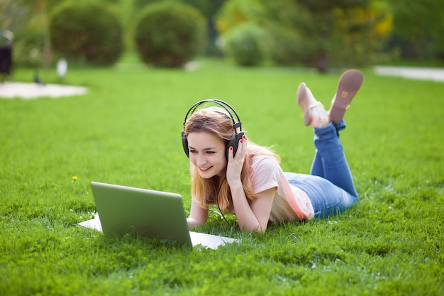 Girl with laptop in earphones lying on the grass