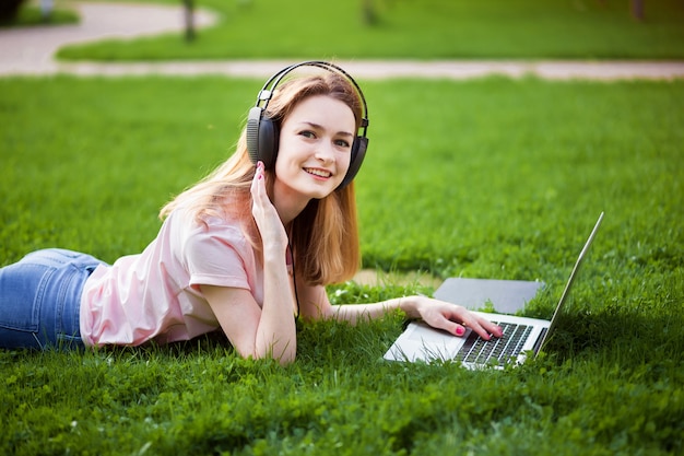 Girl with laptop in earphones lying on the grass