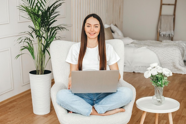 A girl with a laptop sits on a chair looks at the camera and smiles