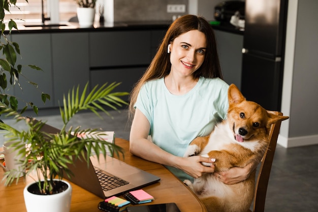 Girl with laptop smile and play with Corgi dog at home Programmer woman working online and have a break for hug her dog Having fun with Welsh Corgi Pembroke