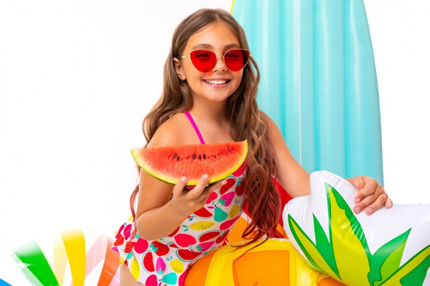 Girl with long crucified hair in sunglasses and swimsuit stands near inflatable mattresses with a watermelon