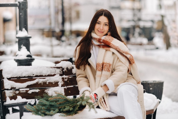 A girl with long hair in winter sits on a bench outside with a bouquet of fresh fir branches