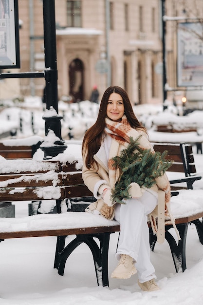 A girl with long hair in winter sits on a bench outside with a bouquet of fresh fir branches