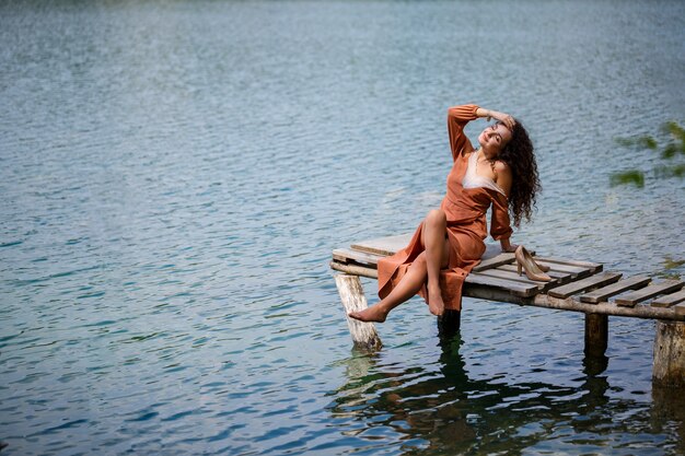 A girl with long wavy curly hair in a long guipure dress barefoot in the summer in a forest on a lake at sunset standing on a pantone on a wooden pier bridge. Summer sunny day