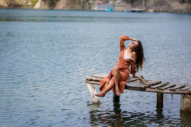 A girl with long wavy curly hair in a long guipure dress barefoot in the summer in a forest on a lake at sunset standing on a pantone on a wooden pier bridge. Summer sunny day