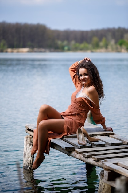 A girl with long wavy curly hair in a long guipure dress barefoot in the summer in a forest on a lake at sunset standing on a pantone on a wooden pier bridge. Summer sunny day